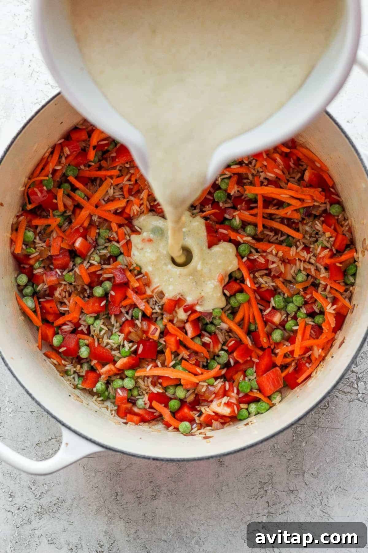 Rich coconut curry sauce being poured over sautéed onions, bell peppers, carrots, and peas in a Dutch oven.
