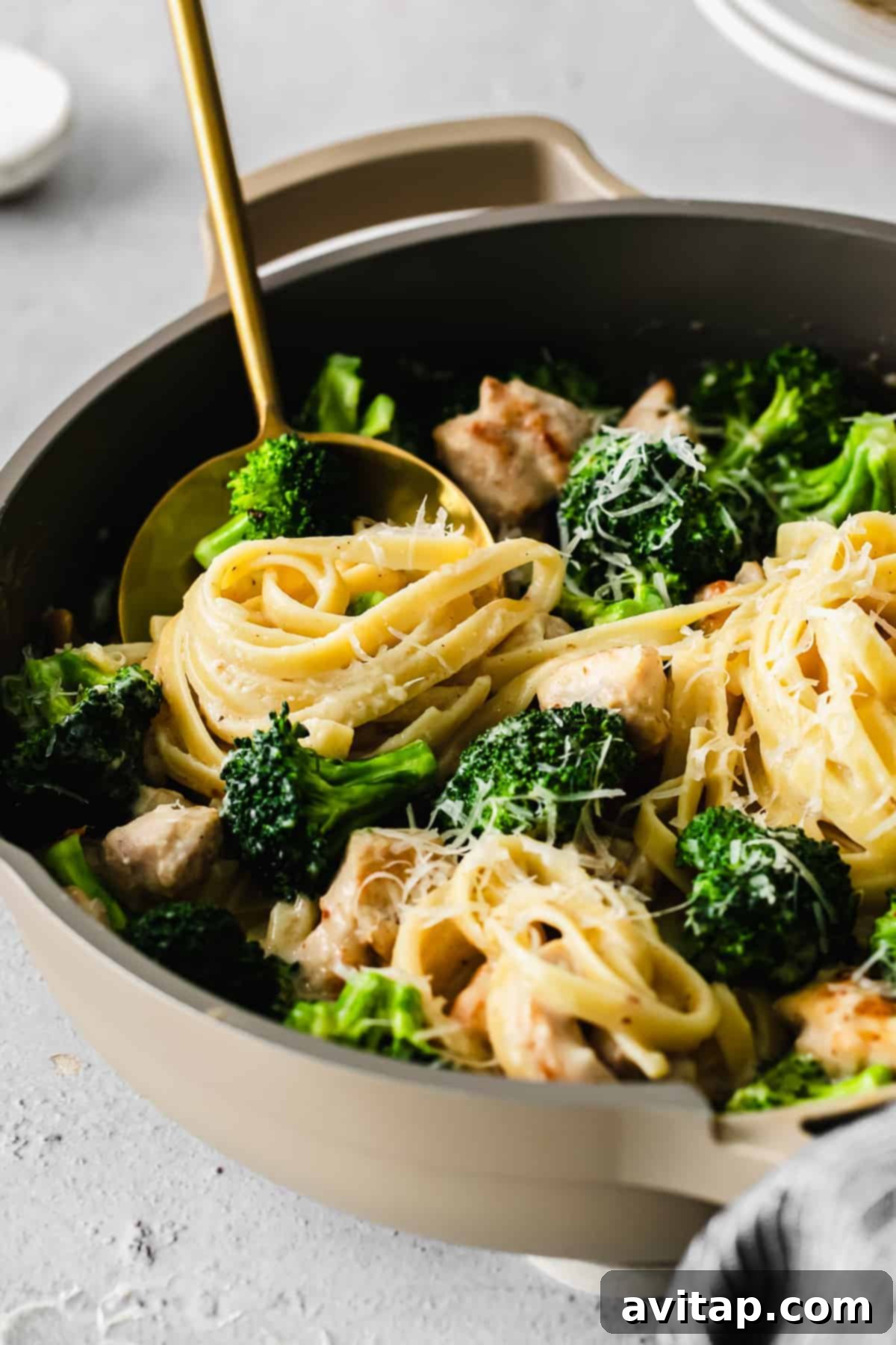 A close-up of a spoon lifting fettuccini noodles and broccoli from a creamy chicken alfredo dish.