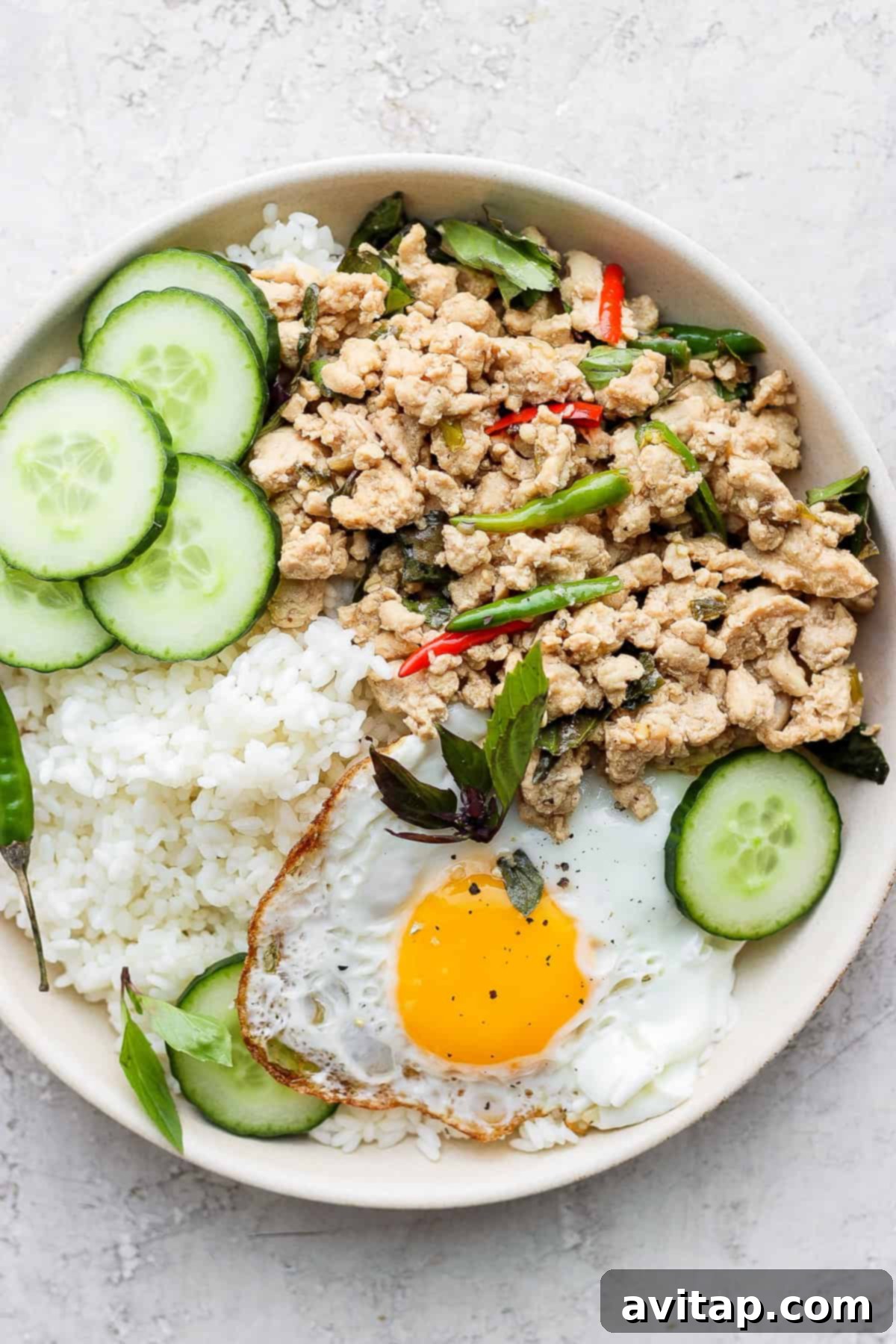 Pad Krapow Bowl with Fried Egg and Sticky Rice A tantalizing bowl of Pad Krapow (Thai Basil Chicken Stir Fry) served with sticky rice, fresh cucumber slices, and a perfectly cooked fried egg, creating a complete and balanced meal.
