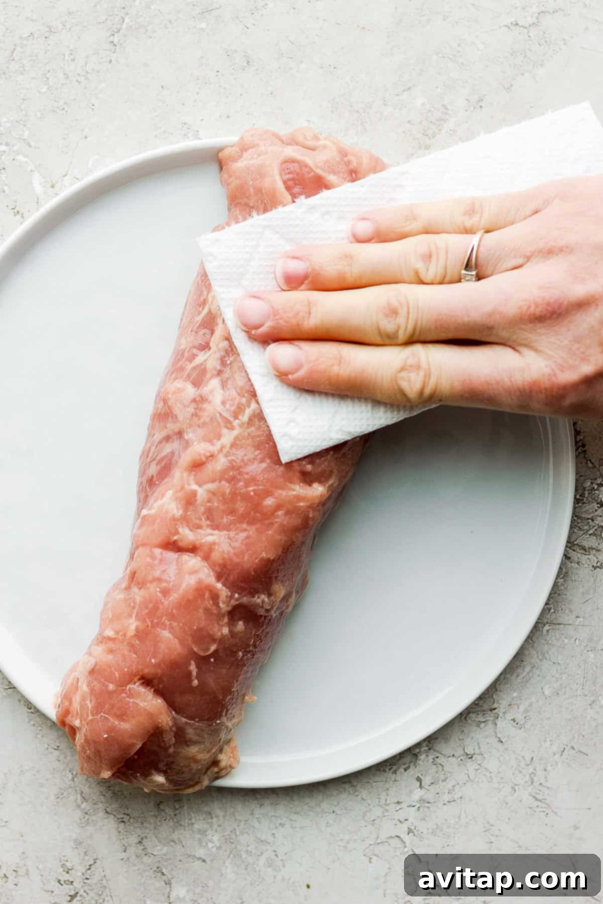 Pork tenderloin being patted dry with a paper towel on a white plate, a crucial step for achieving a crispy crust.