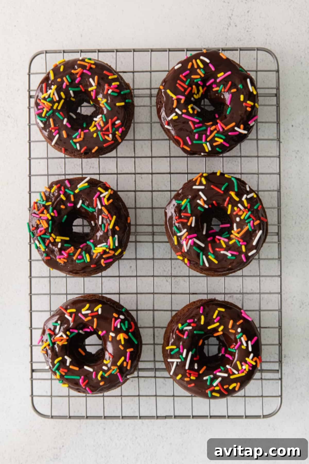 Freshly baked chocolate donuts resting on a wire rack after being glazed and decorated with sprinkles.