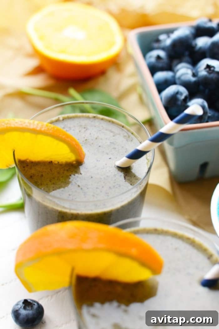Two vitamin-packed green smoothies in glass cups sitting on a wooden table, garnished with orange slices.