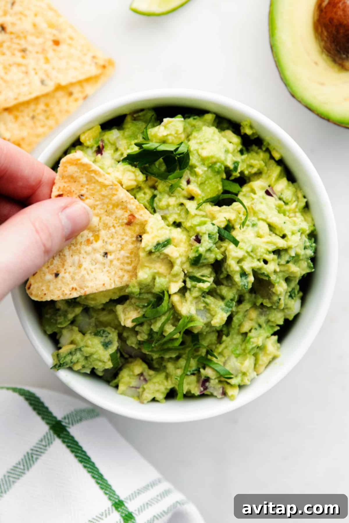 A hand dipping a tortilla chip into a bowl of freshly made green guacamole, garnished with cilantro.