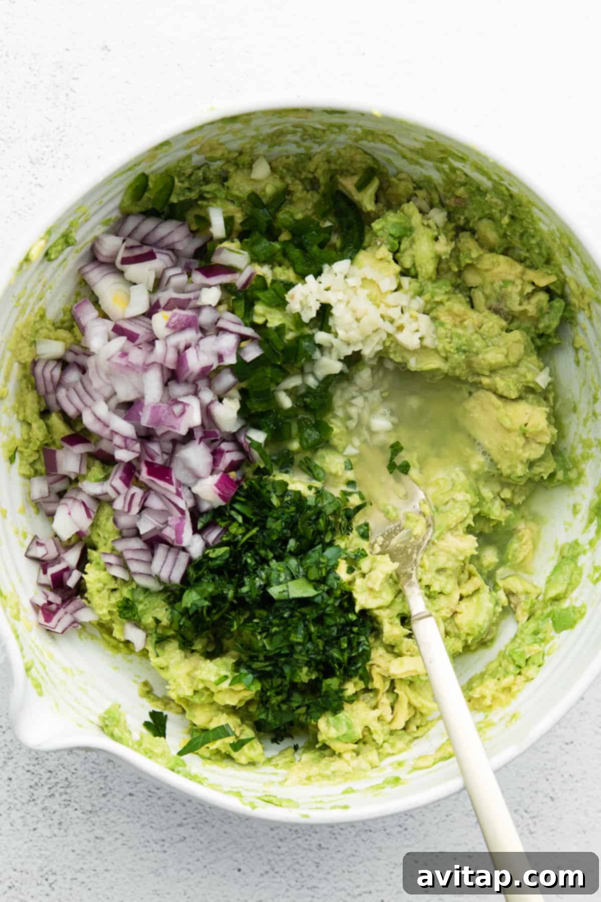 A bowl filled with mashed avocado, cilantro, garlic, red onion, and lime juice, ready to be mixed into guacamole.