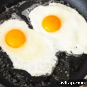 Two eggs are gently frying in a non-stick skillet, with the whites just beginning to set and the yolks still vibrant.