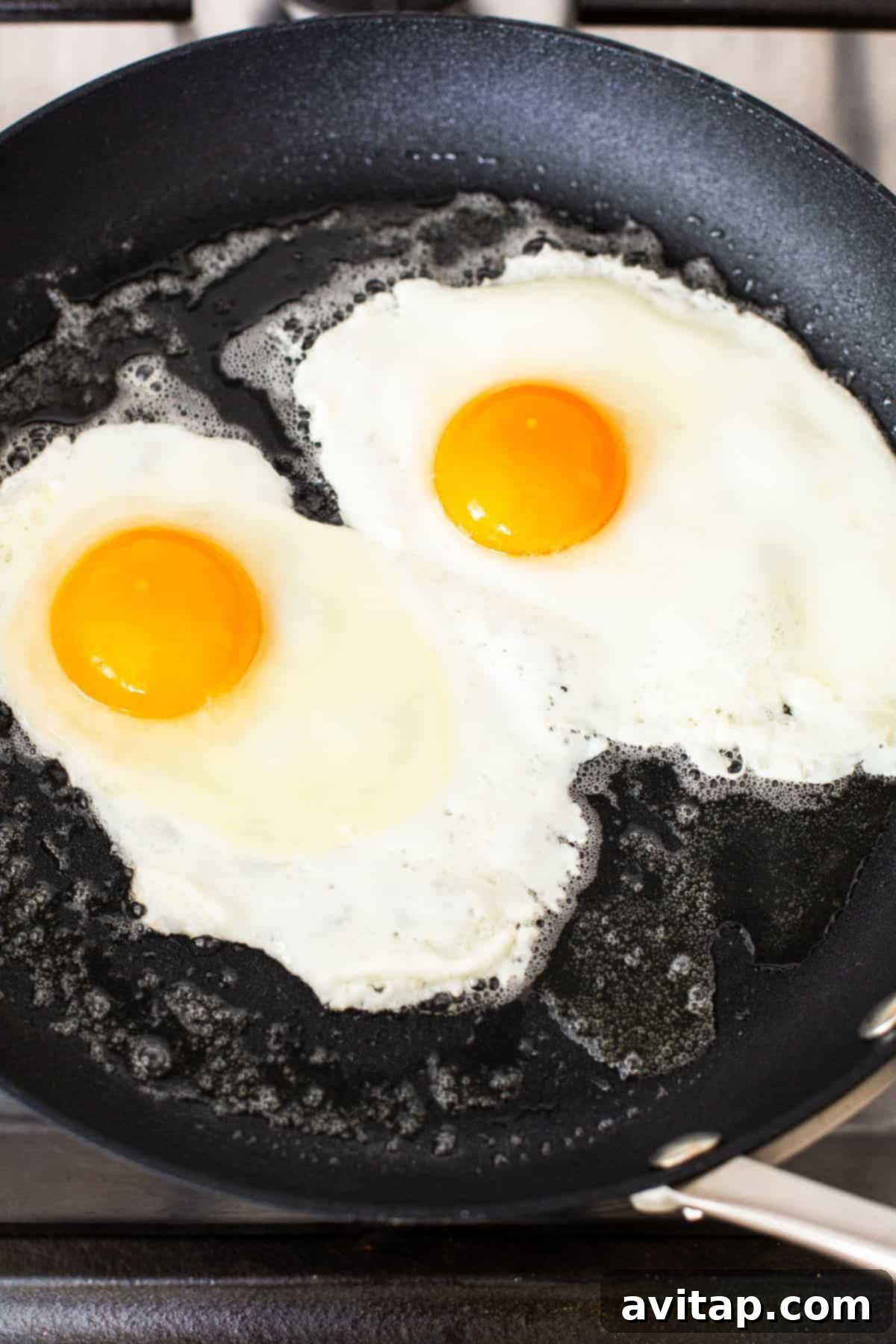 Two eggs are gently frying in a non-stick skillet, with the whites just beginning to set and the yolks still vibrant, on their way to becoming over medium.