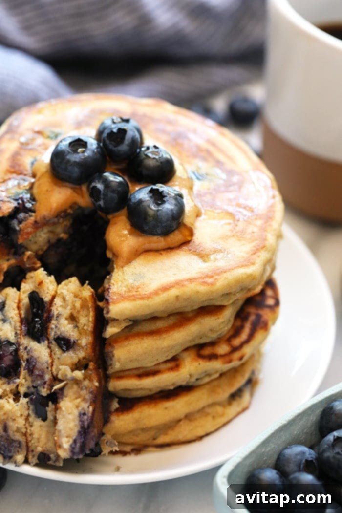 A stack of golden brown blueberry protein pancakes garnished with fresh blueberries and a drizzle of maple syrup, captured from a slightly elevated angle to highlight their fluffy texture and delicious appearance.