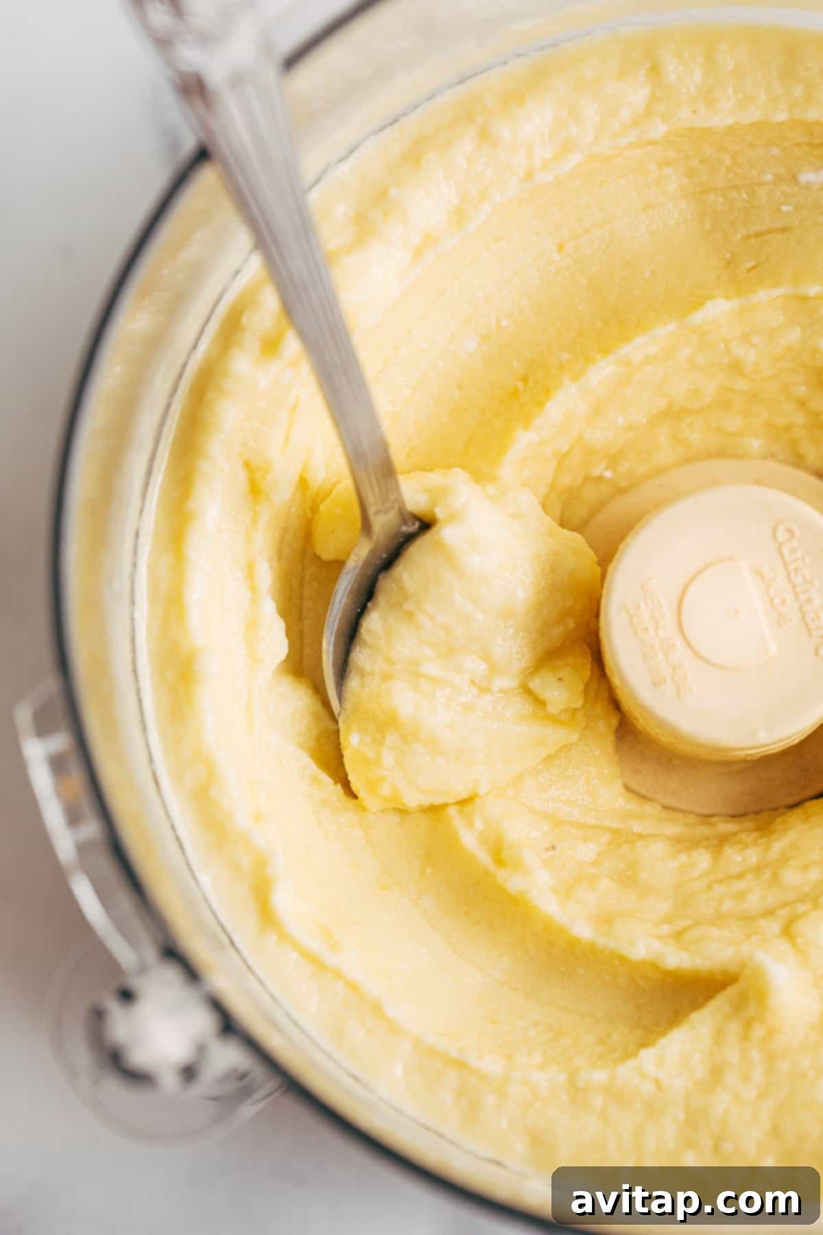 A close-up shot of creamy, yellow Dole Whip being processed in a food processor, with a spoon resting on the side, ready for serving.