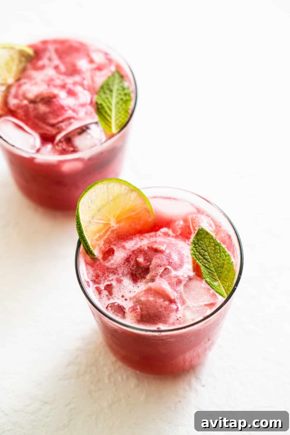 Two glasses of vibrant raspberry vodka punch, garnished with fresh mint and lime, sitting on a table with a pitcher in the background.