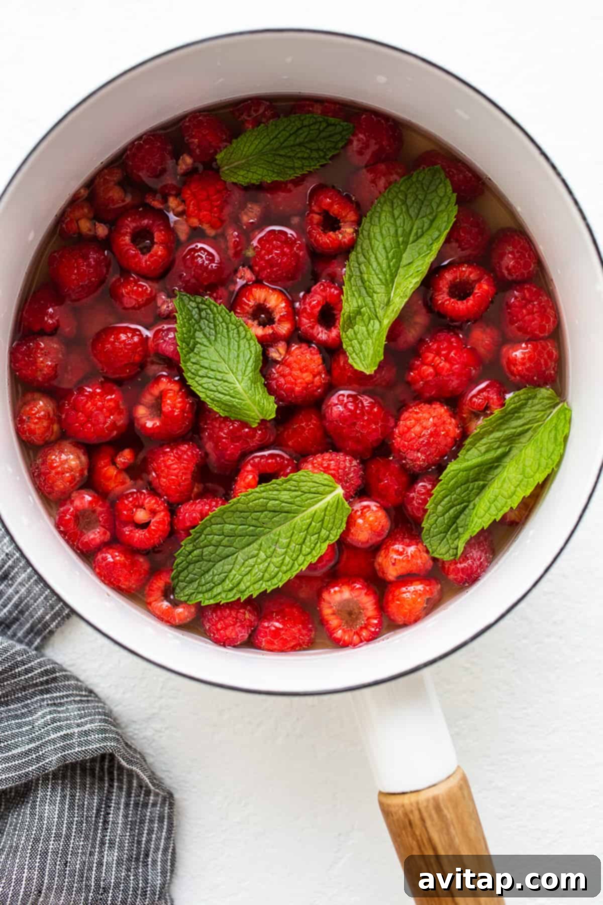 Fresh raspberries simmering gently in a saucepan with water and honey, infused with mint leaves, creating a vibrant red liquid.
