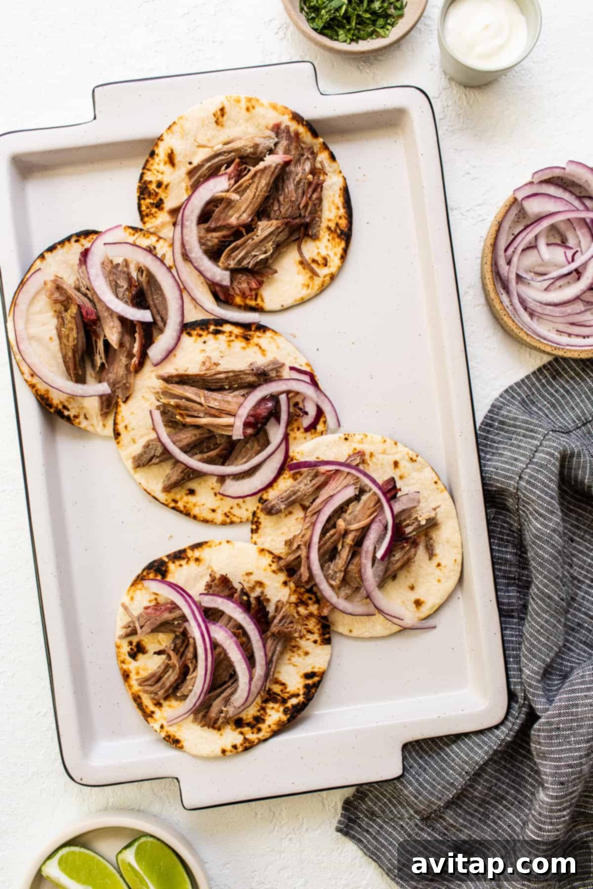 These smoked pulled pork tacos are made with our incredible, fall-off-the-bone smoked pork shoulder and all the best toppings. Partially assembled tortillas with shredded pork and sliced red onion, awaiting more fresh toppings to complete the taco.