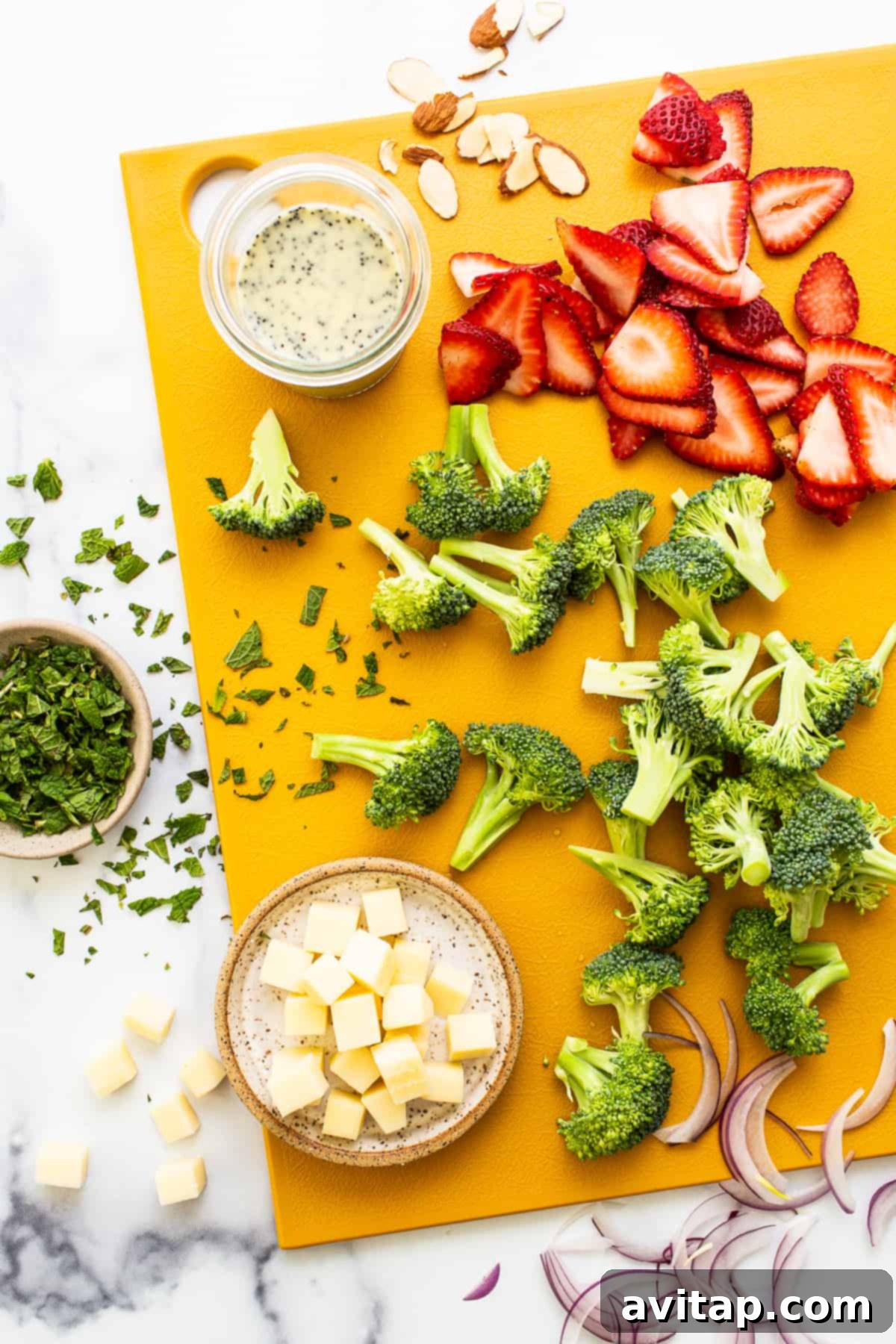 Fresh, colorful ingredients for a homemade strawberry broccoli salad, including broccoli florets, sliced strawberries, red onion, and almonds, beautifully arranged on a wooden cutting board.