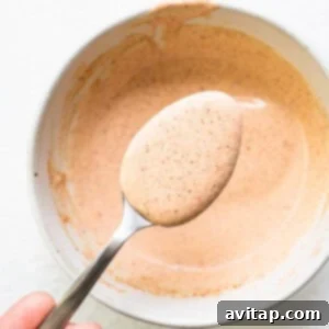 A person holding a spoon full of a creamy, rich brown burger sauce in a small, rustic ceramic bowl, presented beautifully and ready to be served alongside a gourmet burger.