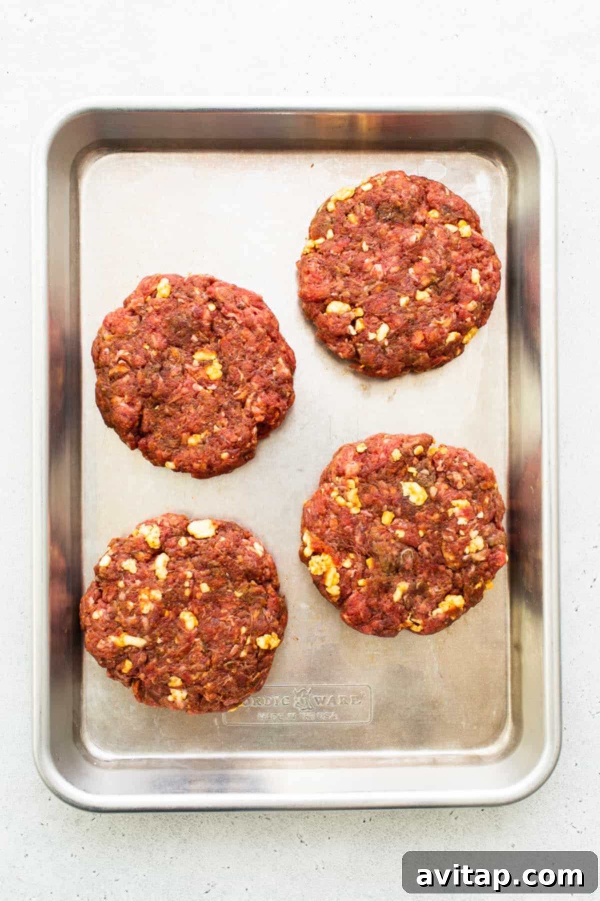 Four perfectly formed homemade burger patties, each featuring a subtle dimple in the center, neatly arranged on a clean baking sheet, perfectly prepped and ready for the cooking process.