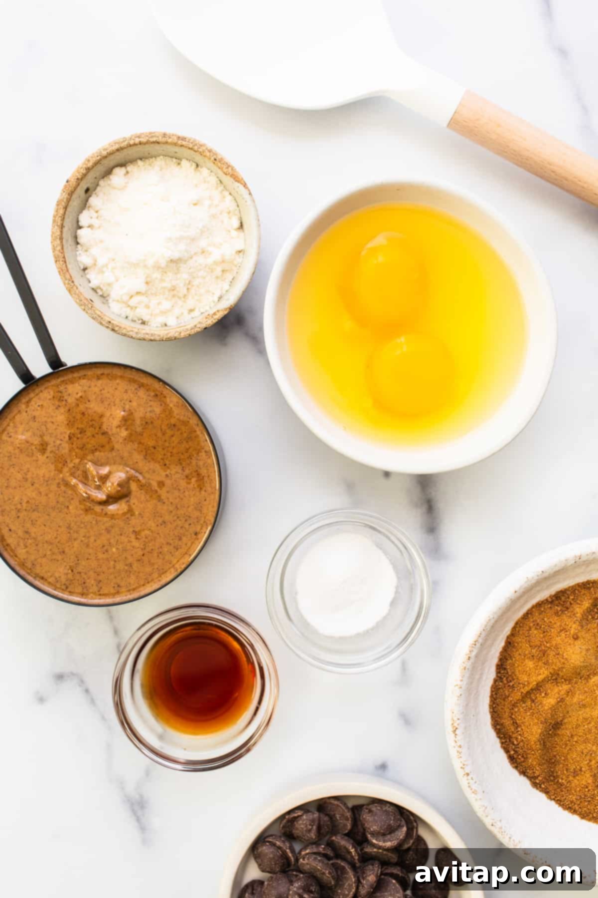 A collection of essential ingredients for making almond butter cookie bars, including almond butter, eggs, coconut sugar, coconut flour, vanilla extract, and chocolate chips, neatly arranged on a kitchen counter.