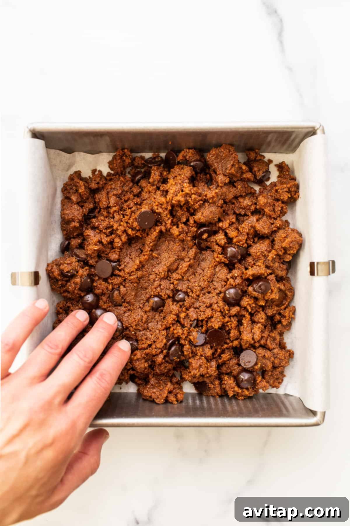Hands pressing cookie dough evenly into a baking pan lined with parchment paper, preparing for baking.