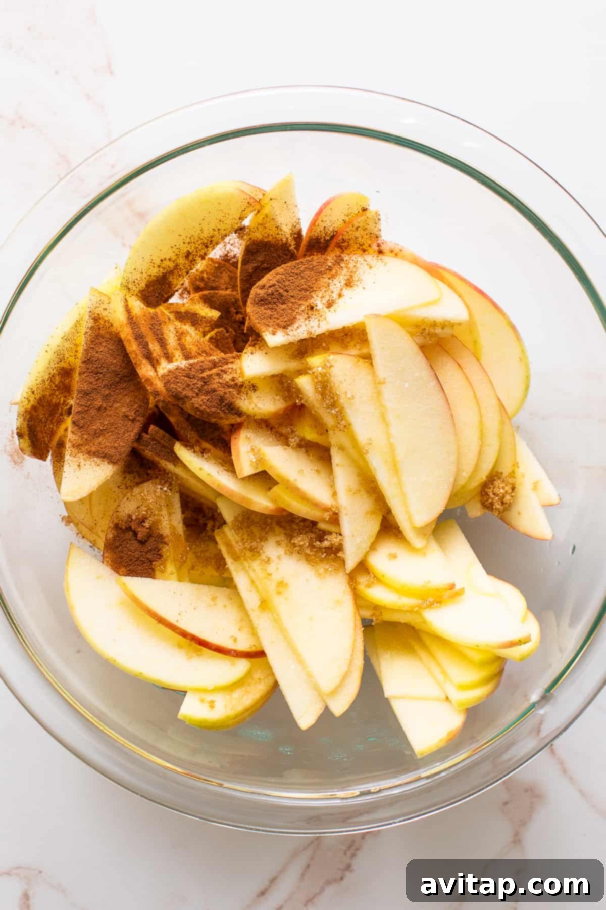 A glass bowl filled with thinly sliced apples, lightly coated with cinnamon and brown sugar, ready to be arranged.