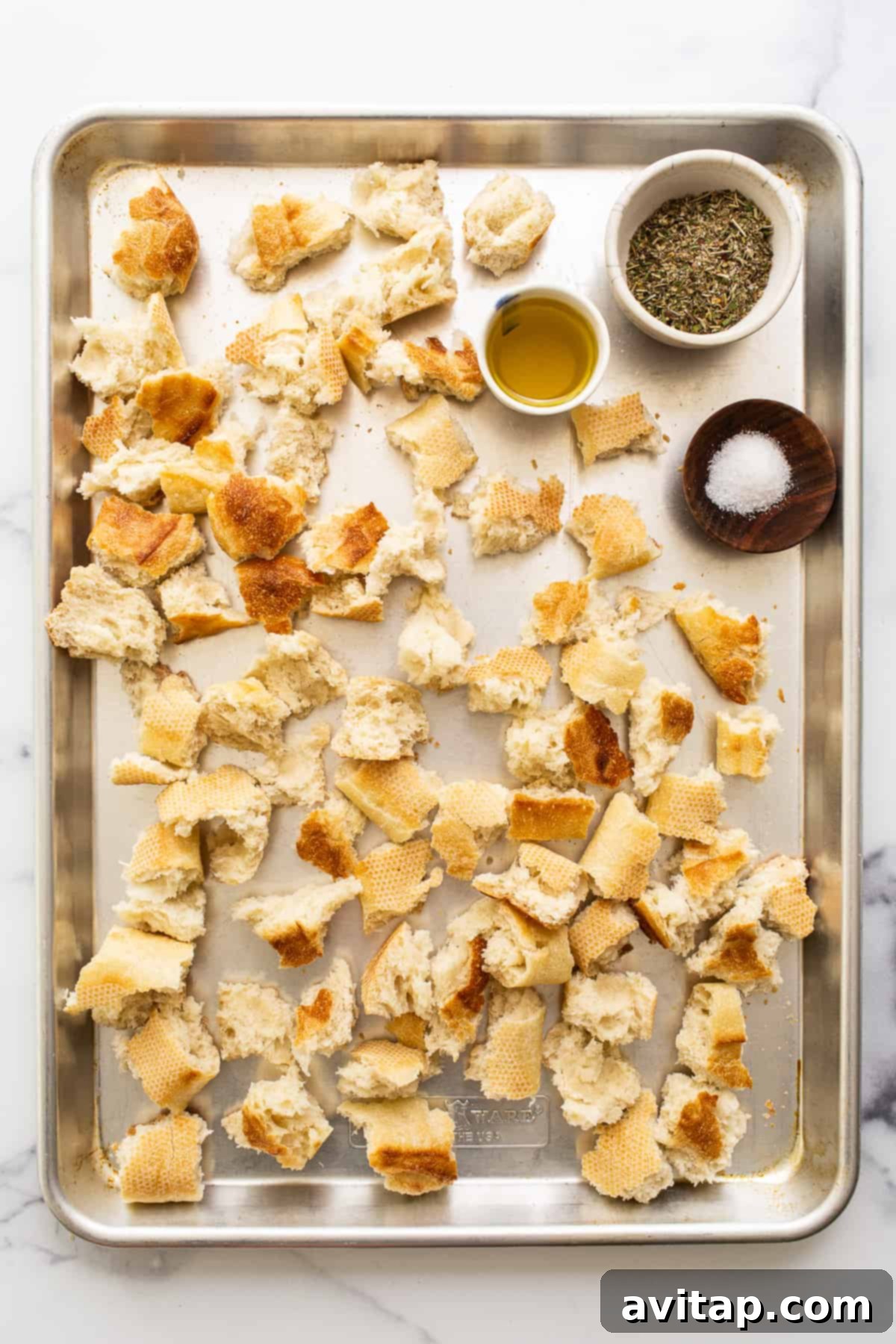 Hand-torn pieces of a French baguette and a small bowl of homemade Italian seasoning displayed on a baking sheet, all prepped for making delicious croutons.