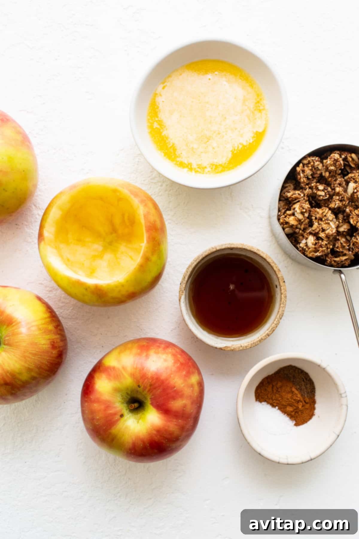 Fresh ingredients laid out for making baked apples: honeycrisp apples, granola, butter, maple syrup, and spices.