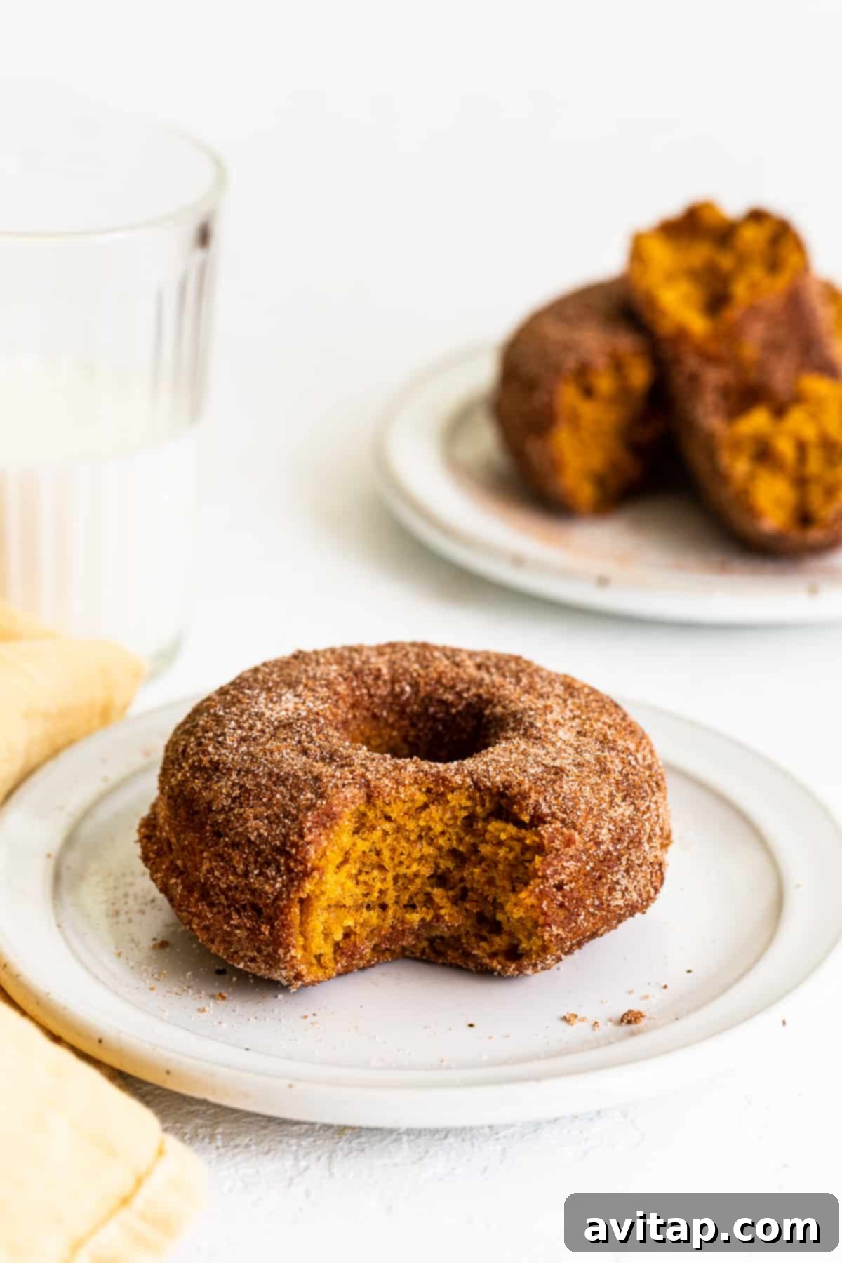 Sink your teeth into these delectable baked pumpkin donuts, featuring real pumpkin and a delightful cinnamon sugar topping. No frying needed! A delectable baked pumpkin donut with a bite taken out, resting on a rustic white plate, coated in sparkling cinnamon sugar.