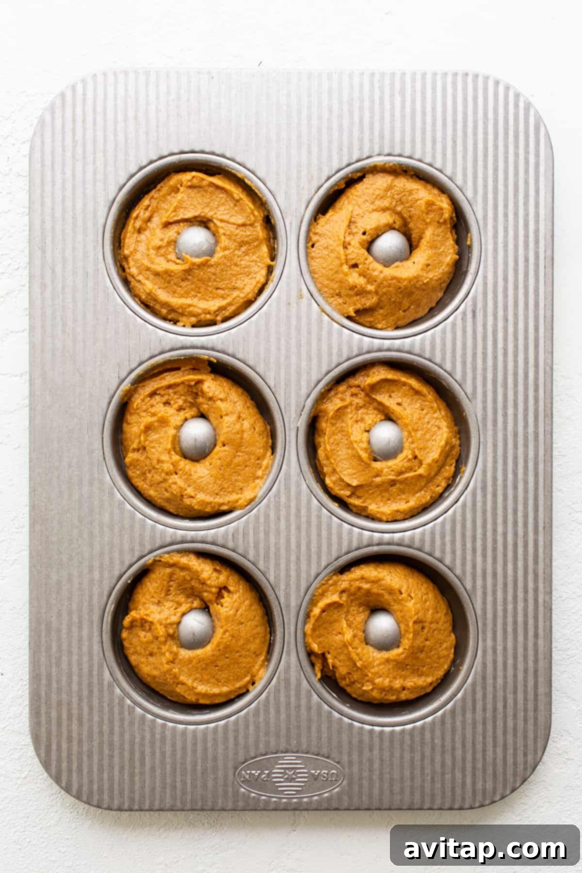 Piping the perfect amount of batter into a donut pan for beautifully baked pumpkin donuts. A donut pan being carefully filled with smooth pumpkin donut batter using a piping bag, illustrating the preparation step before baking.