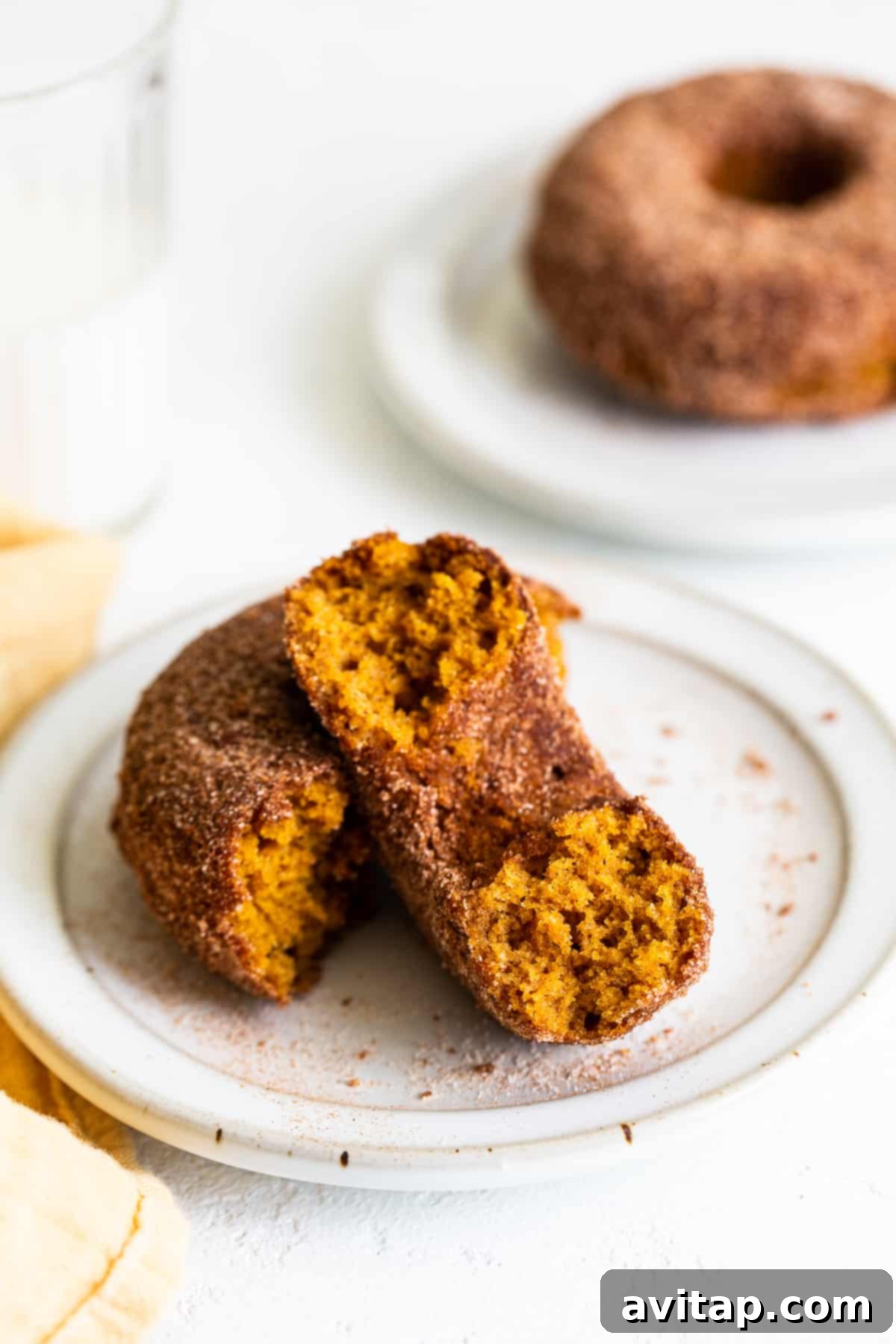 Ready to enjoy: perfectly baked pumpkin donuts, glistening with cinnamon sugar and inviting you to take a bite. A close-up shot of three perfectly golden-brown baked pumpkin donuts, generously coated in sparkling cinnamon sugar, artfully arranged on a pristine white plate beside a small fork.