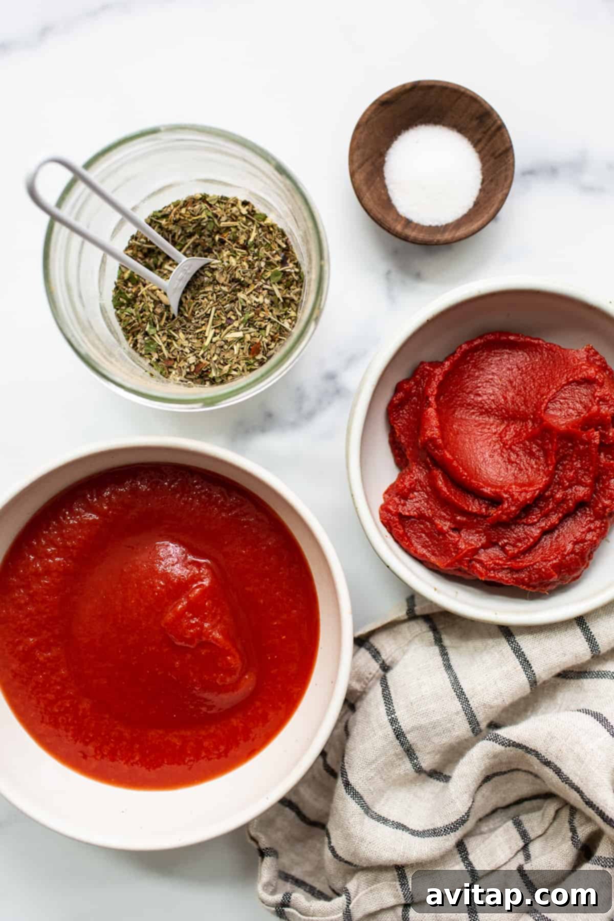 Four individual jars holding the simple ingredients for homemade pizza sauce: canned tomato sauce, tomato paste, Italian seasoning, and granulated sugar.