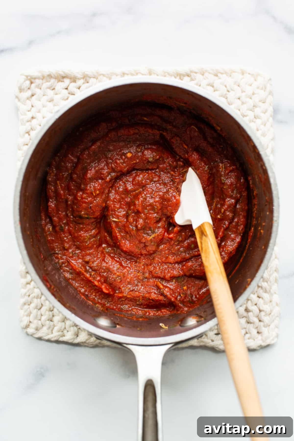 Homemade pizza sauce gently simmering in a small saucepan, with a wooden spoon resting inside.