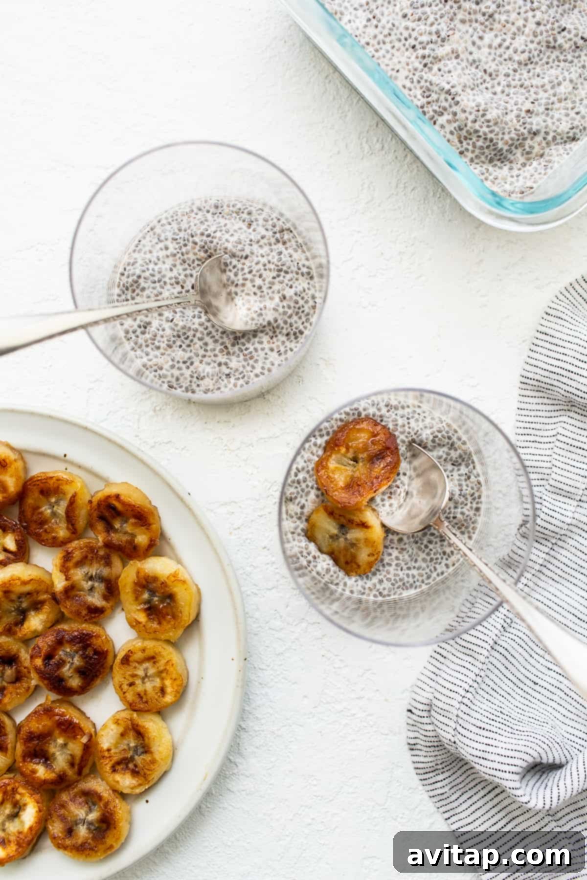 Four individual servings of Banana Cream Pie Chia Seed Pudding in small glasses, garnished with banana slices and a sprinkle of graham crackers.