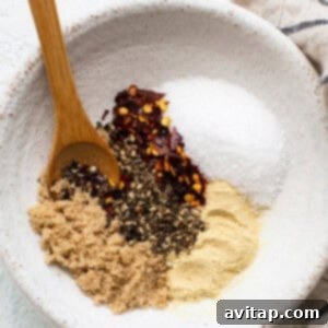 A bowl holds a savory steak seasoning mix of brown sugar, black pepper, crushed red pepper flakes, salt, and garlic powder, with a wooden spoon resting on the side.