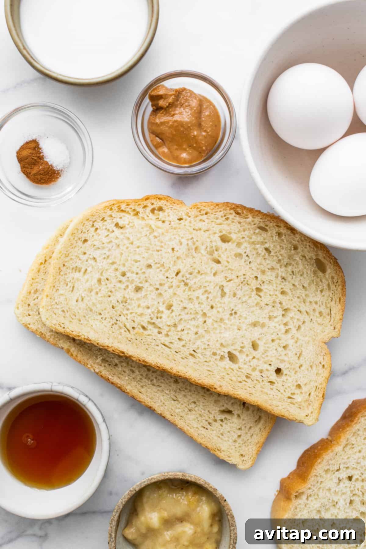 A vibrant selection of ingredients for air fryer chunky monkey french toast including eggs, bananas, milk, maple syrup, peanut butter, cinnamon, and slices of sourdough bread.
