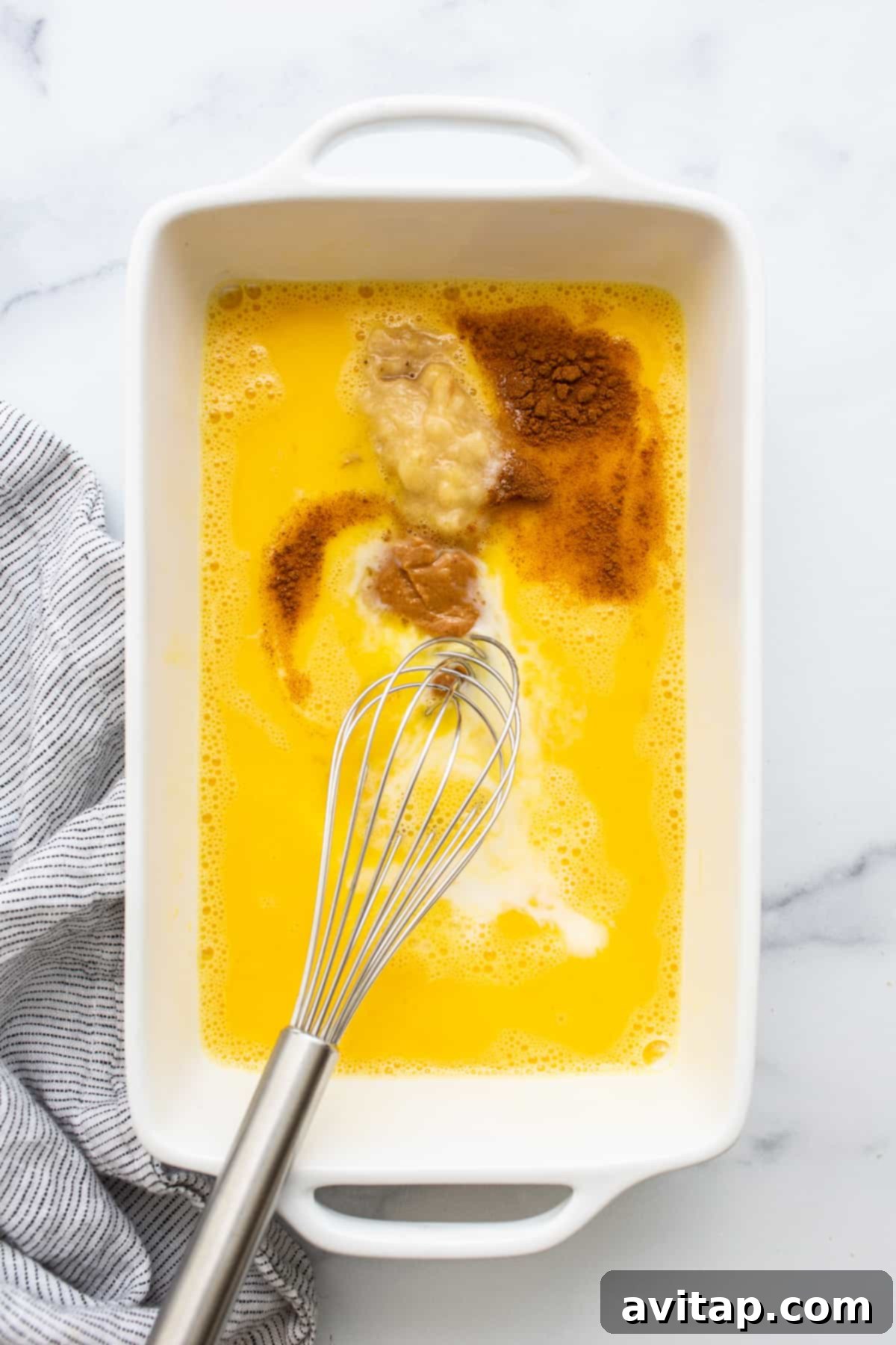 A large bowl filled with the rich egg wash mixture for chunky monkey french toast, with a whisk resting inside, ready for dipping.