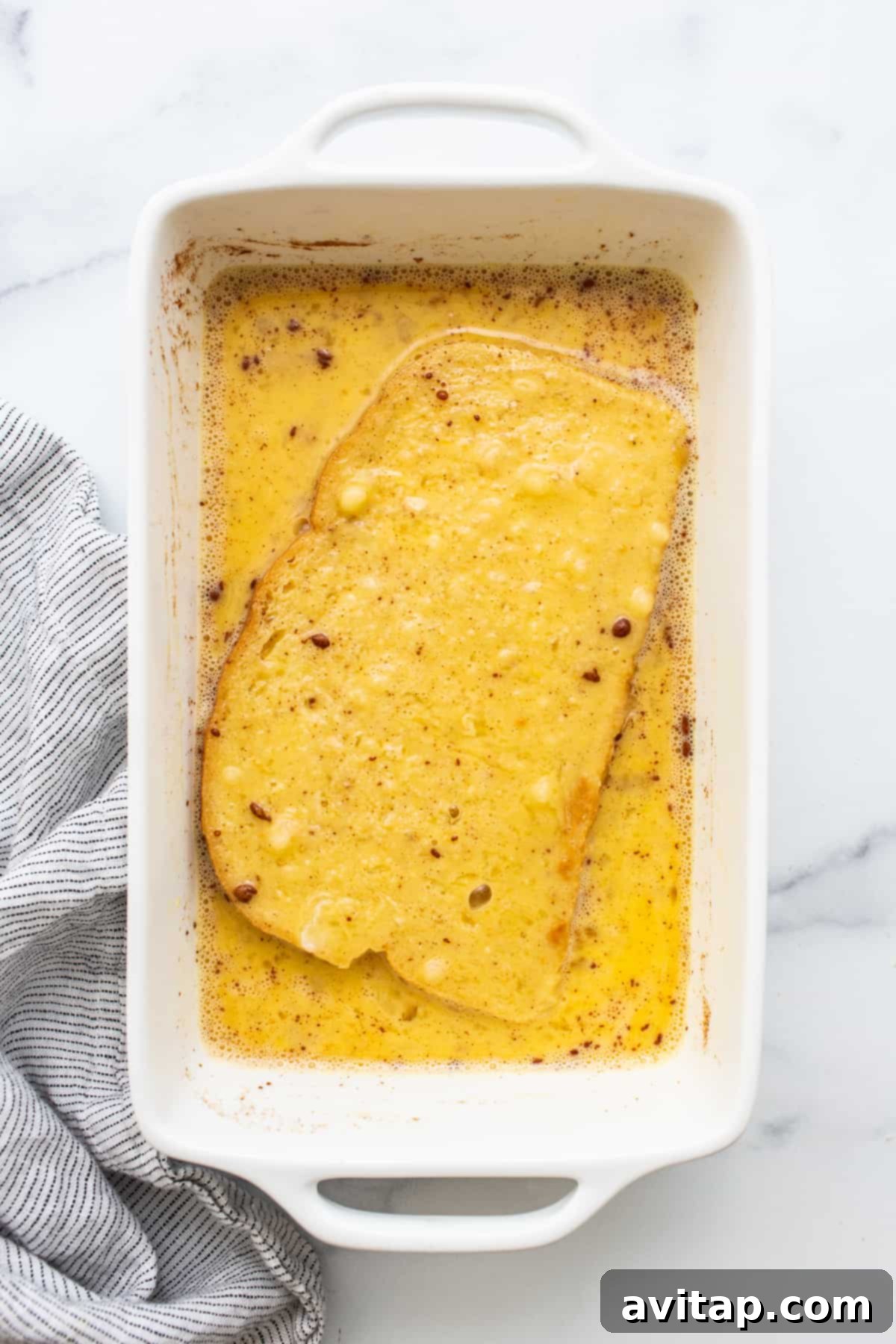 Slices of sourdough bread soaking in a rich, creamy egg wash mixture in a white baking dish.