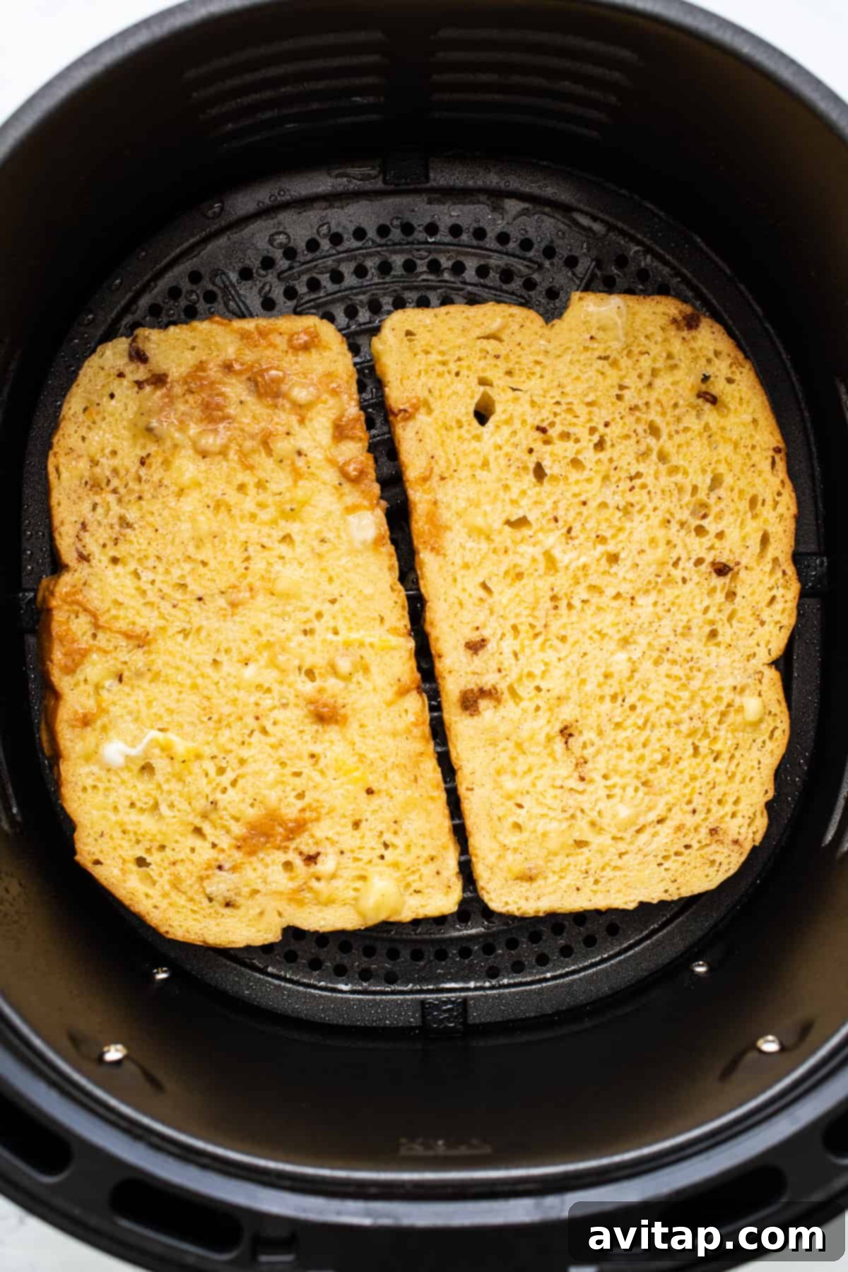 Two slices of golden-brown French toast cooking in an air fryer basket, showing the even crisping achieved by air frying.