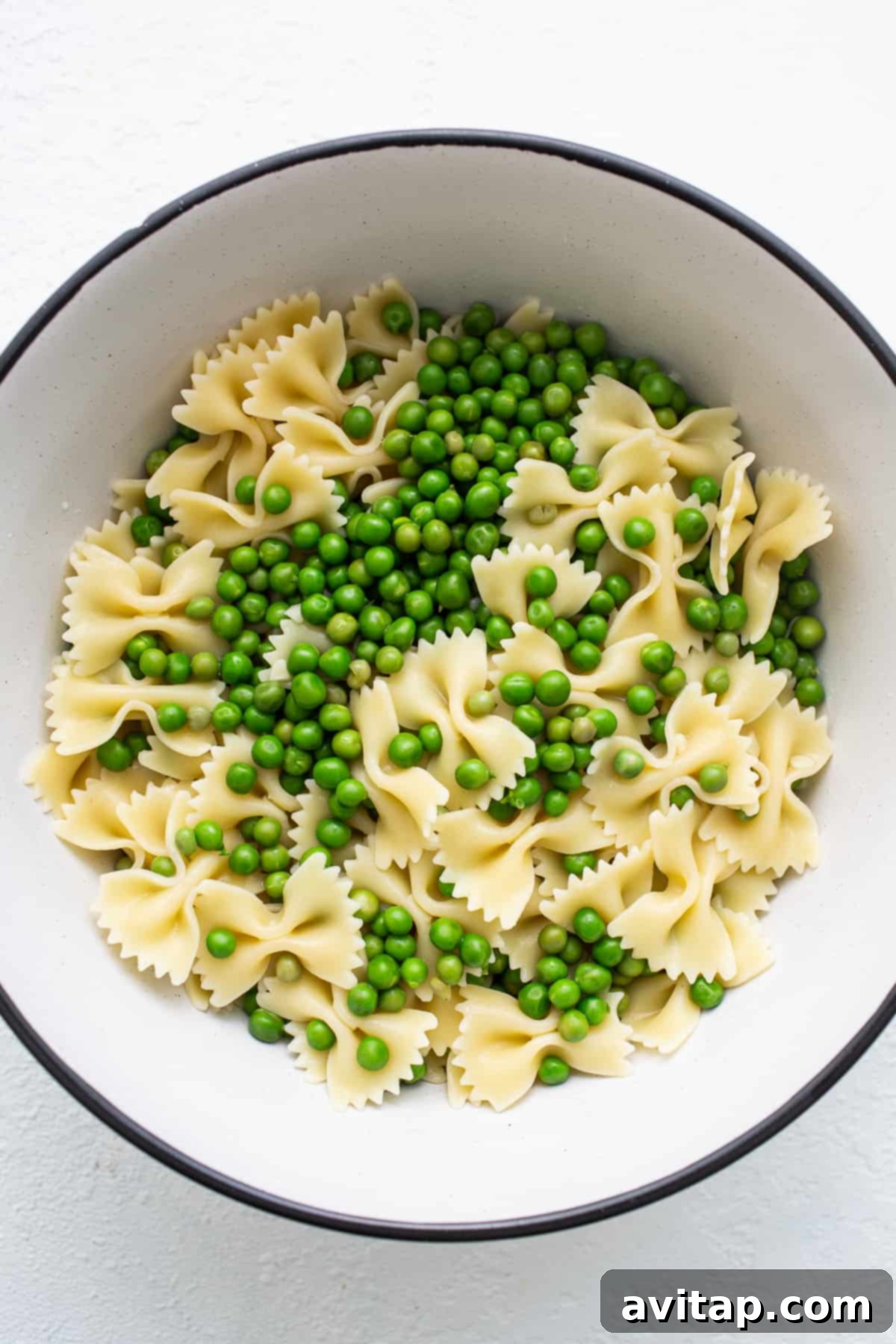 Close-up shot of cooked bow tie pasta mixed with bright green English peas in a white bowl, ready for other ingredients.