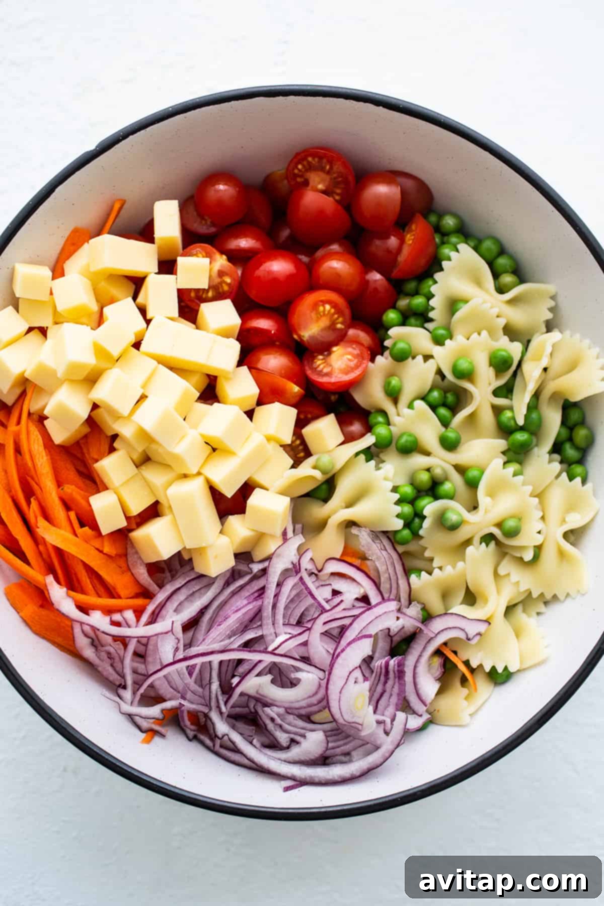 Various fresh ingredients for bow tie pasta salad, including halved cherry tomatoes, sliced red onion, carrot ribbons, and white cheddar chunks, arranged in a bowl.