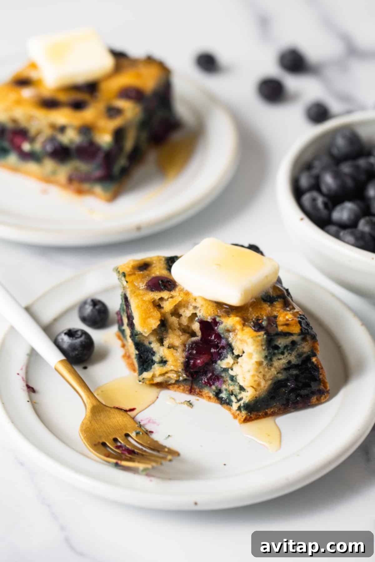 A square of moist blueberry breakfast cake being carefully lifted from the baking pan using parchment paper, showcasing its perfect shape and golden crust.
