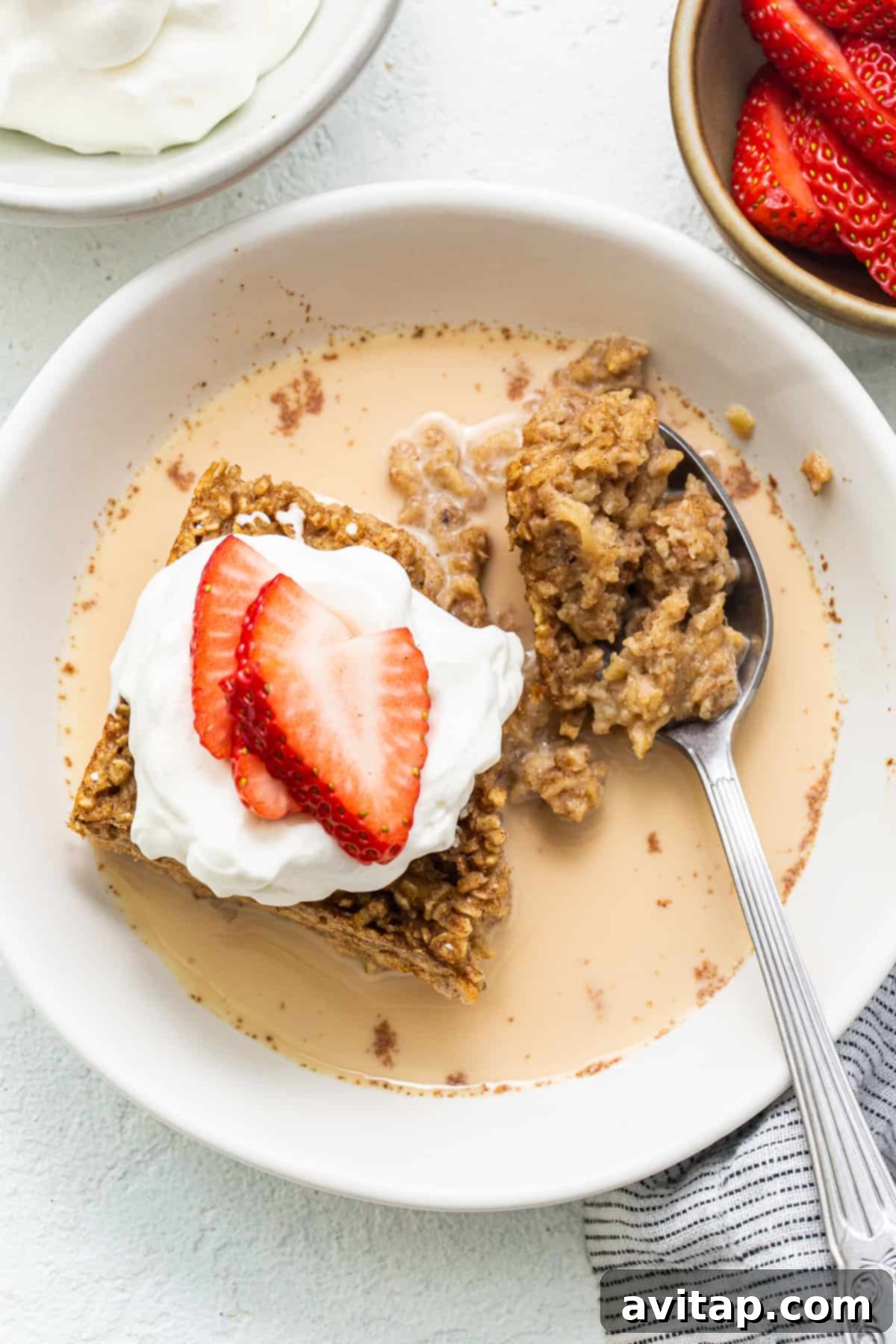 A delectable slice of tres leches baked oatmeal resting in a white bowl, generously topped with vibrant fresh strawberries and a delicate drizzle of the three-milk sauce.