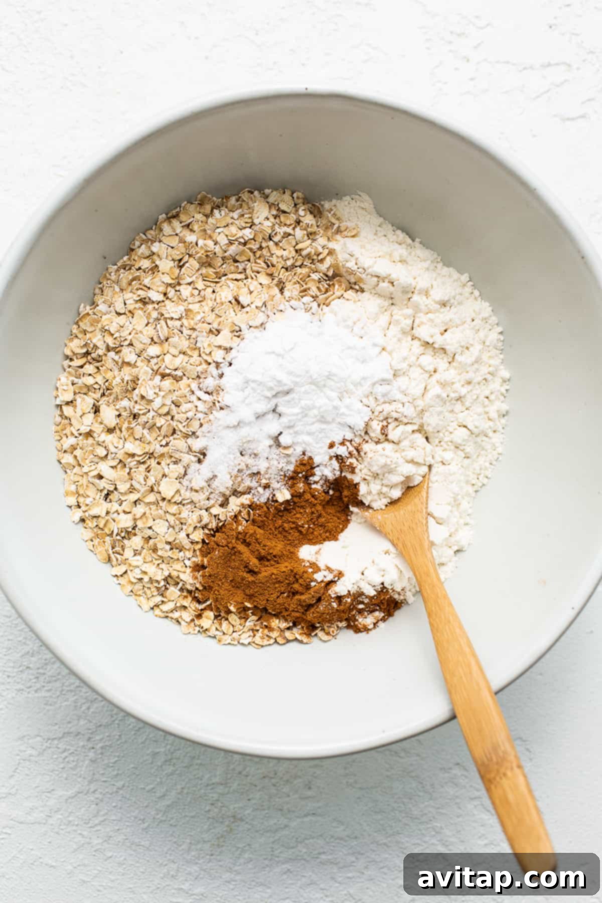 A close-up shot of the carefully measured dry ingredients for baked oatmeal—quick oats, flour, and cinnamon—neatly arranged in a large mixing bowl, alongside a wooden spoon, ready for combination.
