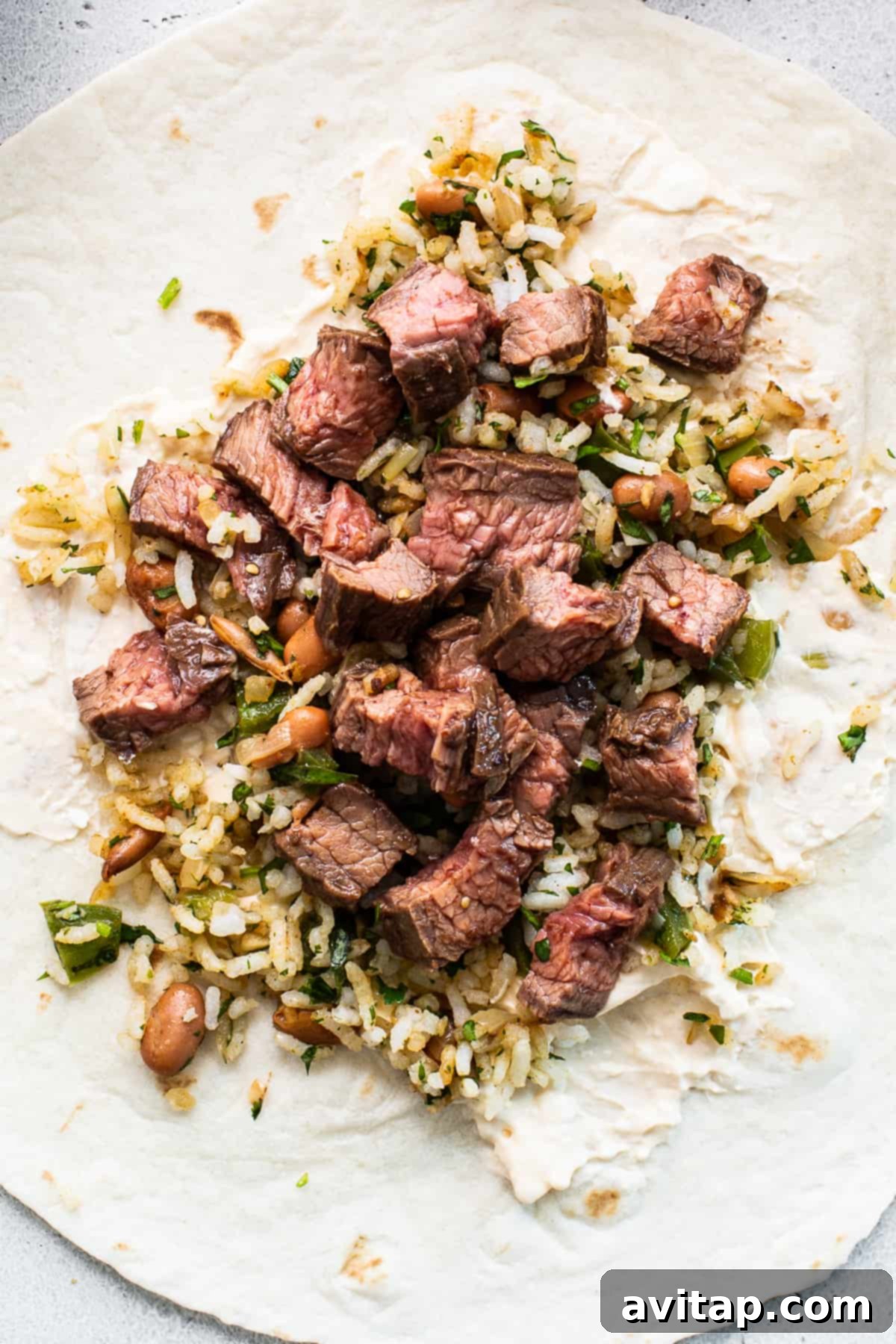 A large flour tortilla filled with perfectly cooked rice and a generous portion of diced marinated steak, awaiting the final roll-up on a light wooden cutting board.