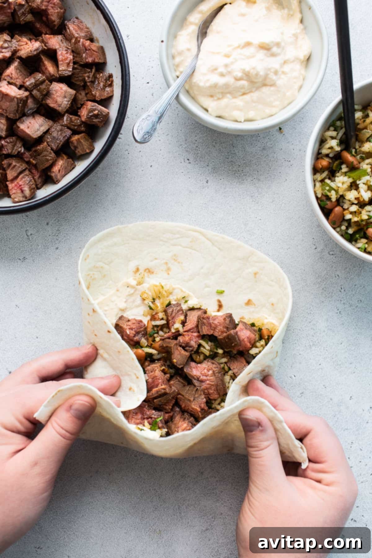 A pair of hands skillfully rolling a steak burrito on a clean kitchen counter, showcasing the assembly process and the generous filling.