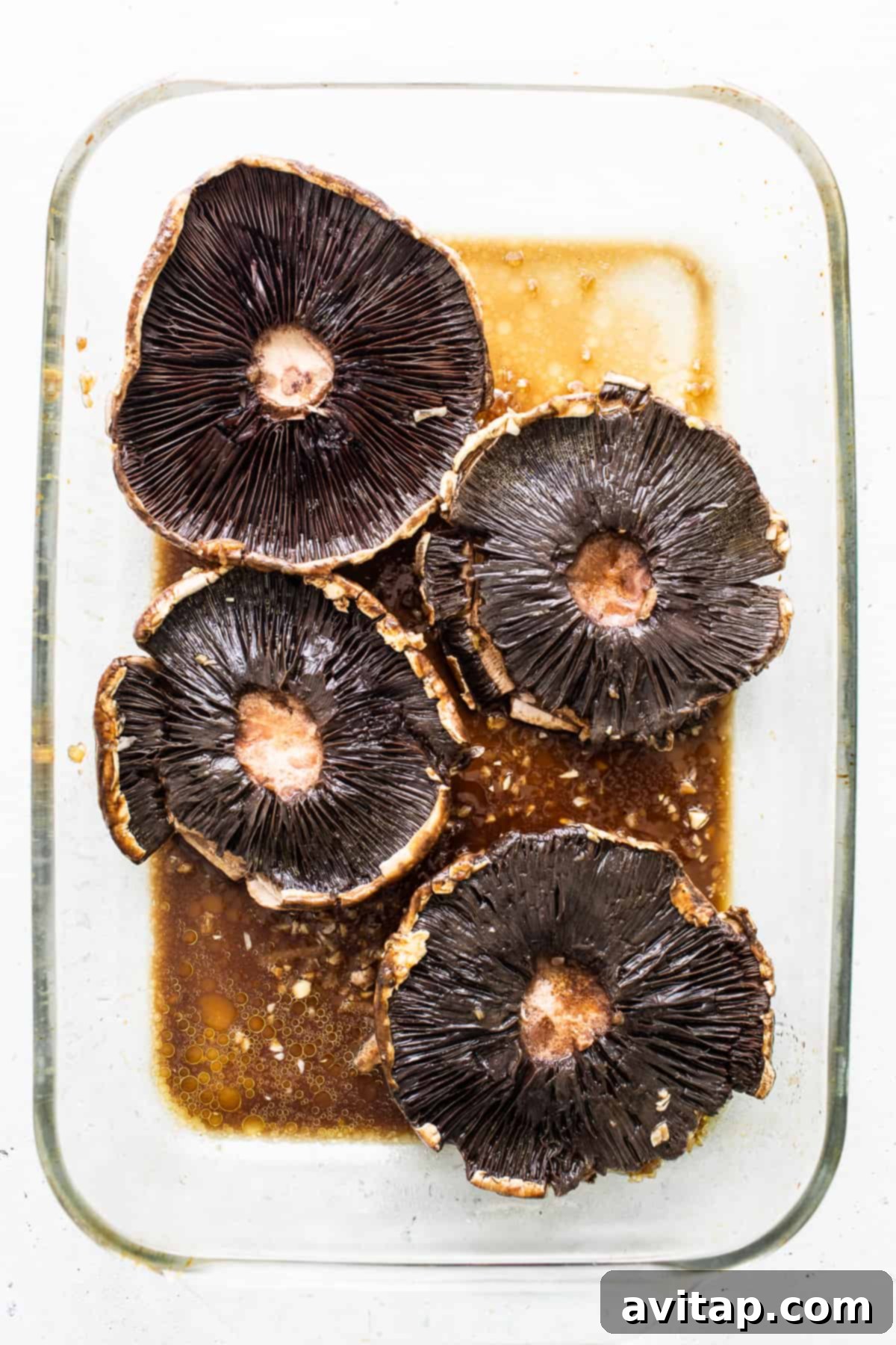 Large portobello mushroom caps soaking in a rich, dark marinade in a glass bowl.