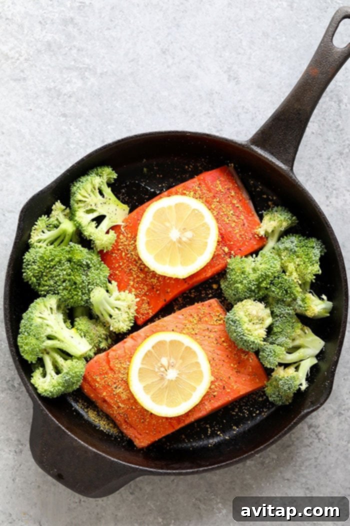 Succulent lemon pepper salmon fillets and tender-crisp broccoli florets baking in a cast iron skillet with fresh lemon wedges.