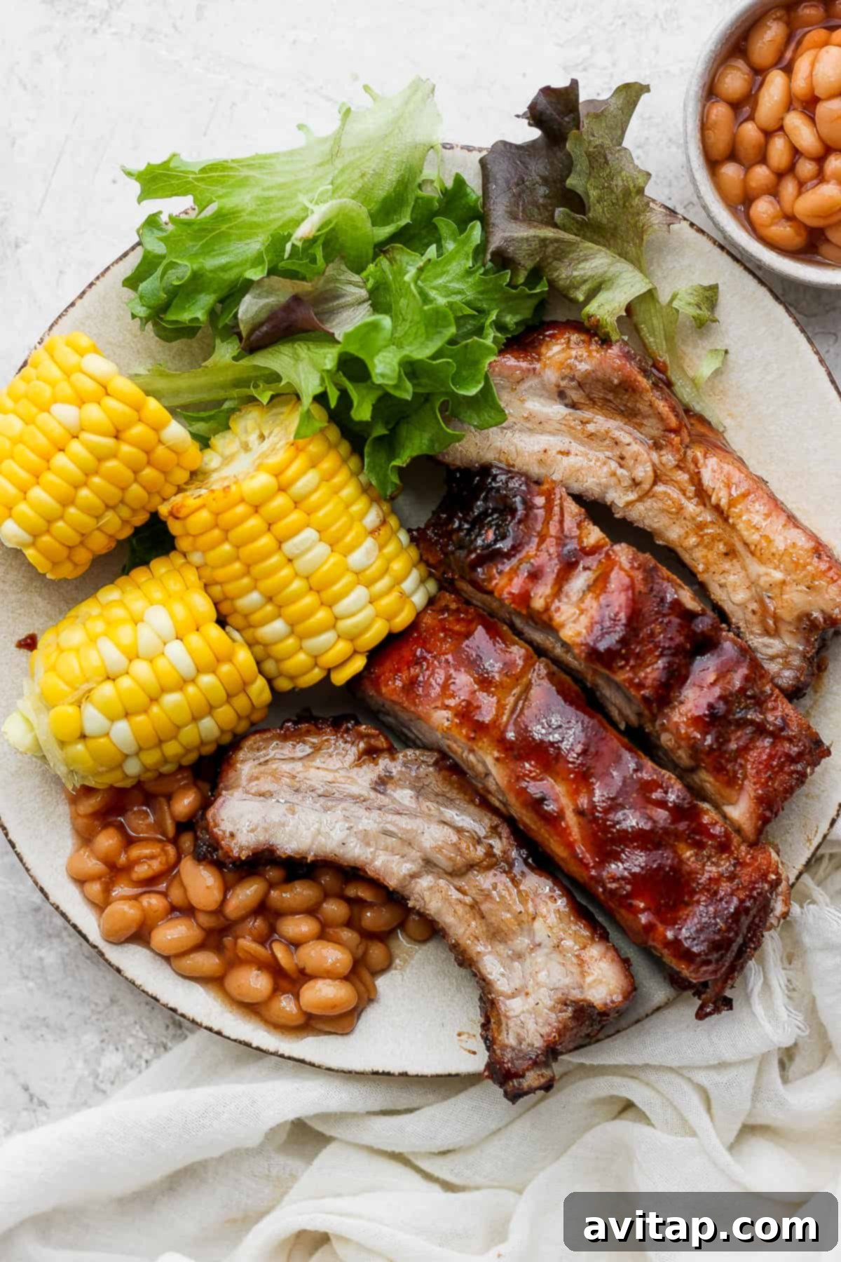 A plate featuring perfectly cooked baby back ribs, served alongside corn on the cob, green beans, and a fresh lettuce salad.