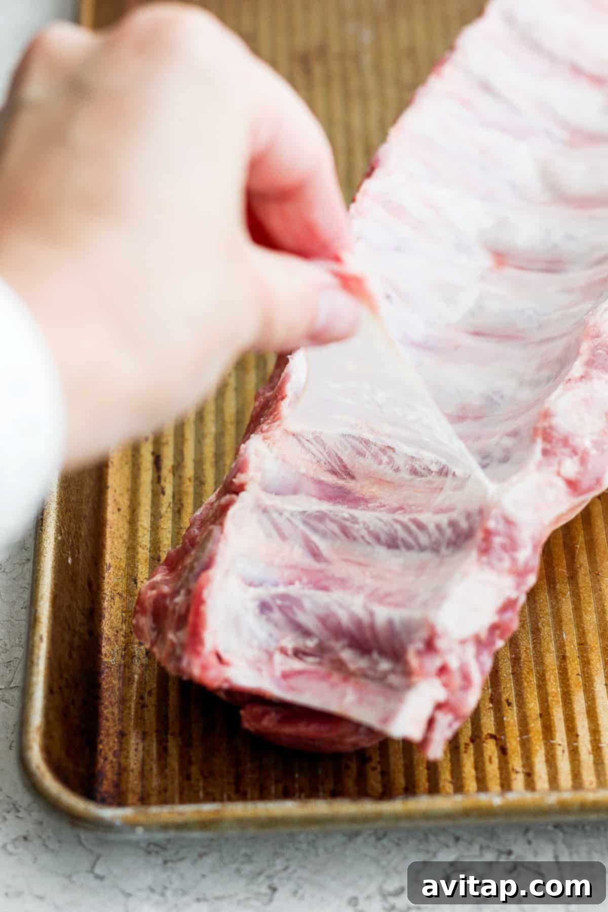 A person is holding a large rack of marinated baby back ribs on a tray, ready for the next step in the cooking process.