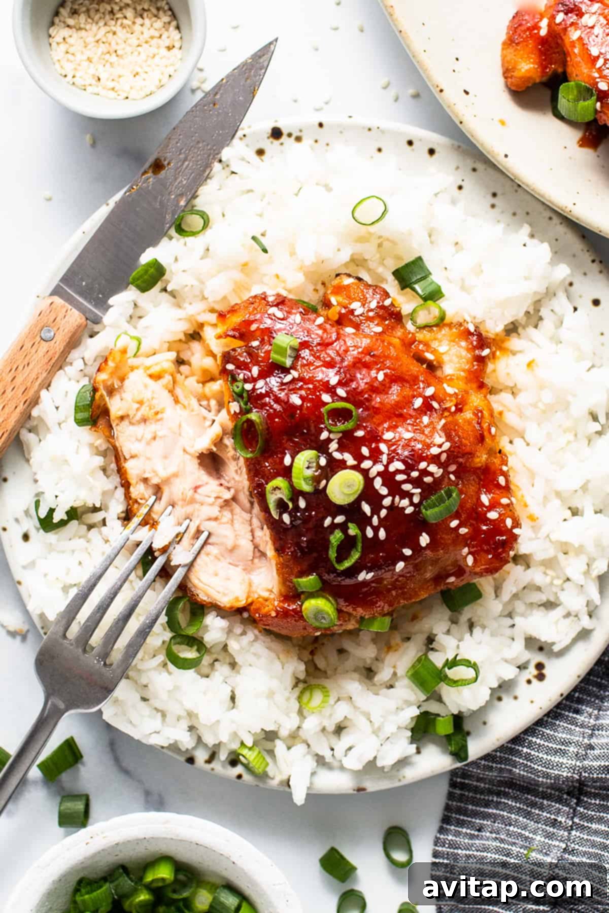 Crispy firecracker chicken thigh on a plate with steamed white rice and a fork, garnished with fresh green onions and sesame seeds.