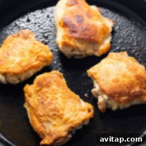Four pieces of seasoned chicken thighs searing in a hot cast iron skillet, bubbling in olive oil to create a crispy crust.