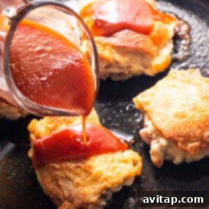 A hand pouring a rich red firecracker sauce over seared chicken thighs in a skillet, creating a sizzling effect.