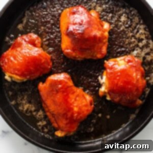 Four saucy chicken thighs resting in a cast iron skillet on a marble countertop, coated in firecracker sauce and ready for baking.