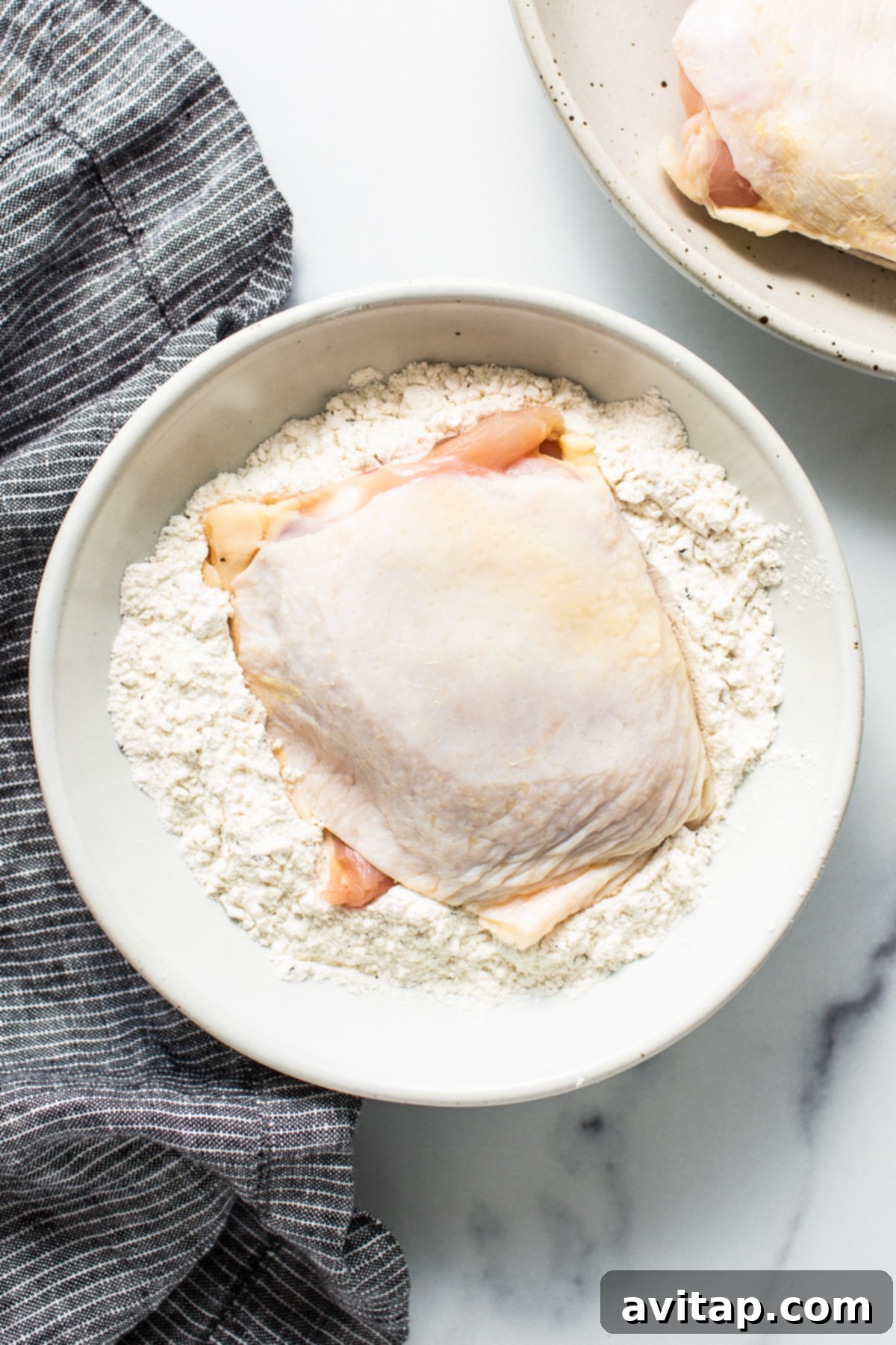 Raw chicken thigh being dredged in a bowl of seasoned flour mixture, ensuring an even coating.